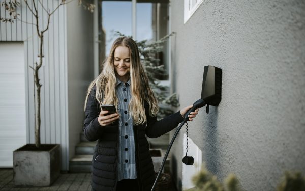 Dame som står foran inngangspartiet til et hvitt hus med glassdør og setter i elbilladeren i veggen mens hun smiler og ser ned i mobiltelefonen